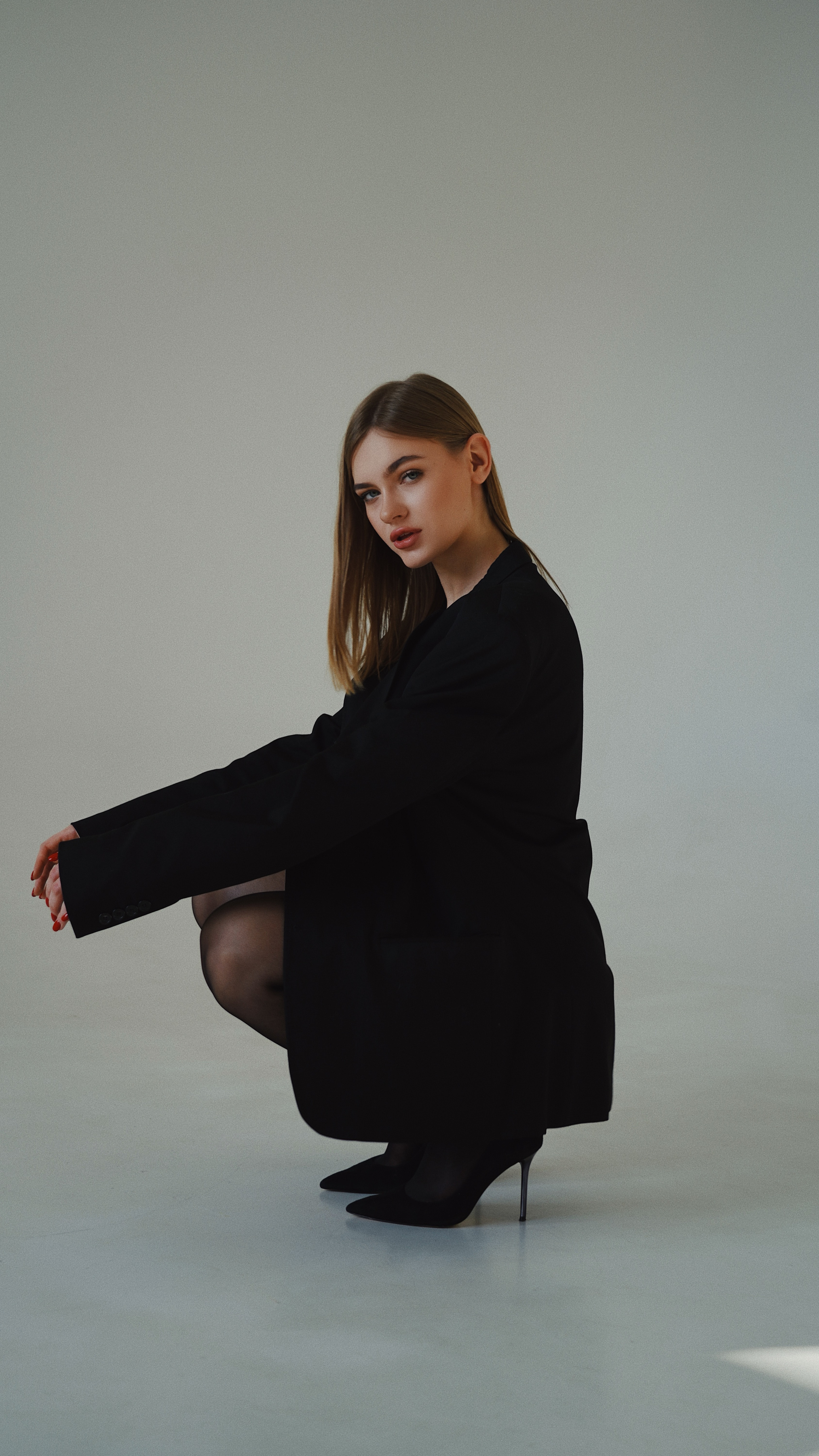Female Model Crouching in Black Jacket and Black Heels 